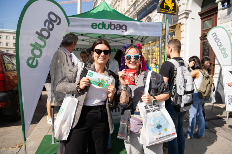 Two women standing in front of an Edubag promotional tent, holding giveaway items and tote bags at an outdoor event