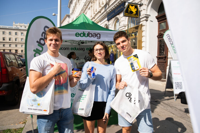 Three young people standing in front of an Edubag promotional stall, each holding branded tote bags and product samples